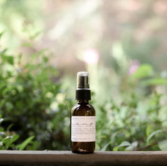 Essential oil and non-toxic Camp Mist Insect Shield by Among the Flowers in amber spray bottle with black lid and white label sitting on wood with green plants in background. 