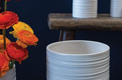 Modern and short striated ceramic Everest Pot in a matte white glaze. Several sizes with red and orange bouquet. Dark navy background. 